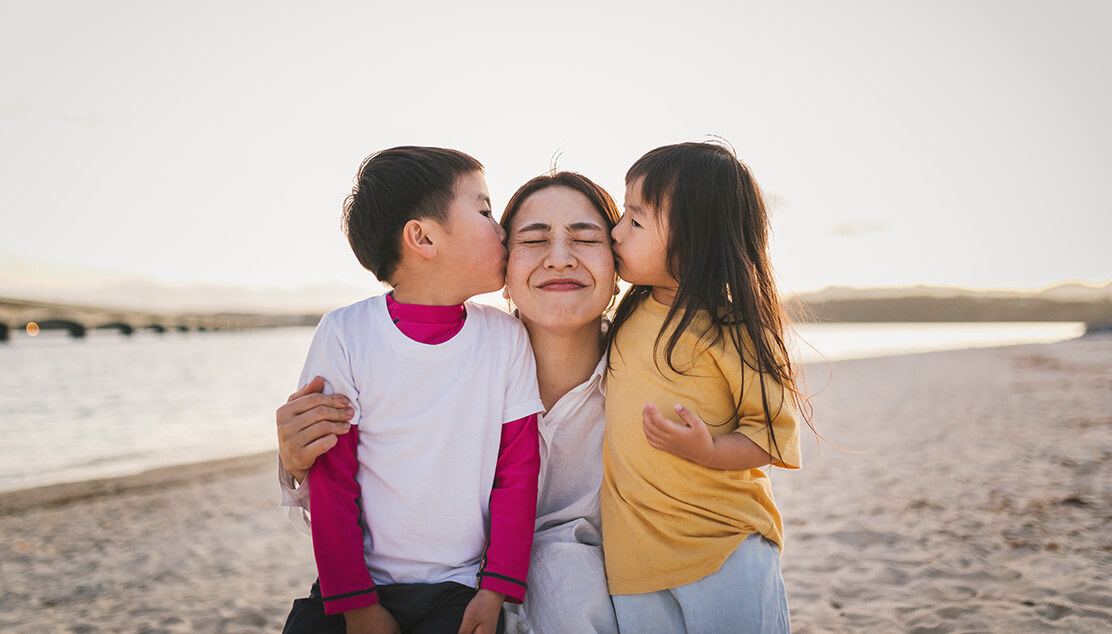 Two children kissing their mother on the cheek at the beach.